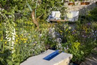 Coin salon avec table et chaises avec coussins, mur en pierres sèches avec nichoirs en acier corten - plantation mixte de plantes vivaces et de fleurs sauvages