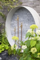 Une pièce d'eau circulaire utilisant des pavés en briques d'argile grises empilés et un contour en béton contre un mur avec des robinets en laiton récupérés comme becs d'eau et un Hortensia arborescens 'Annabelle'.