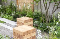 Sièges cubiques en bois dans un jardin de gravier entouré de plantations mixtes de plantes vivaces, dont Delphinium 'Guardian White'