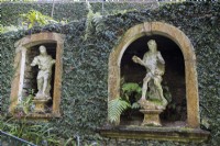 Les vieilles arches de fenêtres en pierre encastrées dans les murs couverts de lierre créent des niches pour les statues figuratives en pierre. Jardins du Monte Palace, Madère. Août. 