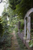 Un chemin en dalles de pierre mène au loin avec des fenêtres cintrées en pierre à côté, au milieu d'une plantation tropicale luxuriante. Jardins du Monte Palace, Madère 