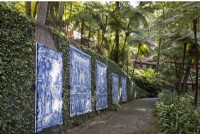 Des panneaux carrelés anciens ornent un mur couvert de lierre avec des arbres au-dessus et un chemin pavé devant. Jardins du Monte Palace, Madère. Août. Été 