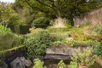 Petit jardin formel avec buis et ifs coupés, un banc rustique et un vieux chêne en toile de fond en octobre 