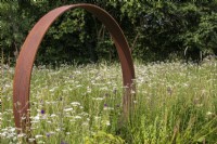 Une porte lunaire en acier Corten se dresse dans une prairie vivace avec Achillea millefolium. 