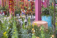 Une vue à travers un parterre de fleurs vivaces jusqu'à un plan d'eau circulaire rose pastel à côté d'un baril de pétrole peint en bleu et blanc planté de fleurs et de légumes dans le jardin d'unité du RHS et de l'Eastern Eye. Créateur : Manoj Malde 