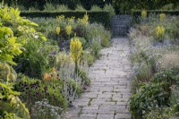 Un chemin pavé mène à travers un parterre de fleurs estival profond avec Achillea millefolium, Allium sphaerocephalon, Kniphofia et Verbascum olympicum 