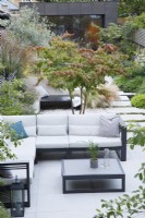 Vue d'ensemble de ce jardin de ville avec le coin salon avec Parrotia persica multi-troncs, chaise longue et chemin menant au bâtiment recouvert de zinc 