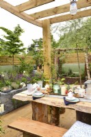 Pergola en bois avec un toit protecteur en plexiglas avec une table dressée dans un jardin d'inspiration boisée entouré d'une clôture en planches de bois. JuinConceptrice : Mary Anne Farenden. Bord Bia Bloom, Super Garden, Dublin, Irlande. 