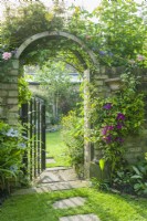 Portail de jardin en fer forgé dans un mur de briques habillé de roses et de clématites. Tremplins menant à travers une pelouse soigneusement tondue. Juin 