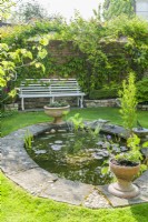 Siège de jardin dans un petit jardin de ville donnant sur un étang ovale avec des nénuphars et des plantes marginales. Juin. 