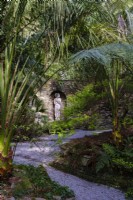 Vue sur le jardin semi-tropical 'secret' avec fougères arborescentes et palmiers, statue classique encastrée dans un mur de pierre 
