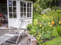 Maison d'été et patio de jardin de campagne avec chaise longue et pots succulents en terre cuite à côté d'un parterre de fleurs mixtes avec Tulipa 'Sunny Prince' et feuillage printanier 