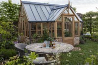 Une salle à manger extérieure avec table recyclée et serre en bois en arrière-plan - 'Manger, boire et être Rosemary' - designer Laura Ashton-Phillips - RHS Malvern Spring Festival 2024 