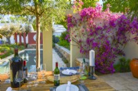 Placez une table en bois dans un patio avec Acacia et Bougainvillea spectabilis en fleurs grimpant sur le mur - donnant sur la piscine dans le jardin méditerranéen. JuinDesigner : Alan Rudden 