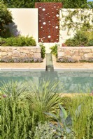 Mur avec panneau en acier avec une cascade d'eau qui se jette dans un ruisseau qui coule jusqu'à la piscine principale et des parterres de fleurs surélevés en pierres avec des plantes exotiques dans le jardin méditerranéen. JuinDesigner : Alan Rudden 